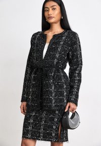 Veste en tweed noir et blanc à manches longues, sans col, ceinturée à la taille. Associée à une jupe crayon assortie. Accessoires incluant un petit sac à main noir.