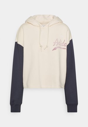 Kreminė trumpa hoodie su tamsiai mėlynomis rankovėmis. Yra raišteliai, siuvinėtas logotipas ir minkšta, tekstūruota medžiaga.