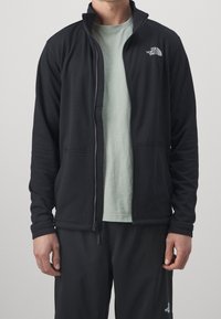 Homme portant une veste noire zippée North Face par-dessus un t-shirt vert clair et un pantalon noir sur un fond gris uni.