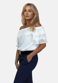 Biała bluzka z odkrytymi ramionami z falbaniastymi rękawami, zestawiona z granatowymi spodniami w kropki, z czarnym paskiem z detalem złotej klamry.