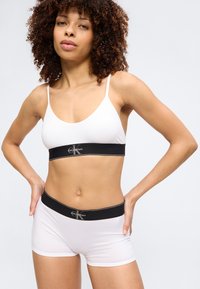 Moteris su garbanotais plaukais, dėvinti baltą Calvin Klein braletę ir šortus su juodais prekės ženklo elastiniais diržais, stovinti rankomis ant klubų.