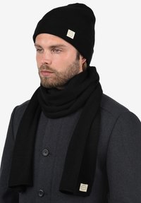 Zwarte geribbelde gebreide beanie en bijpassende sjaal, beide gemaakt van zachte materialen, met een klein logo-label aan het einde van de sjaal.