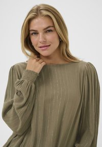 Olivenfarvet bluse med lange, voluminøse ærmer og smockede manchetter. Har vertikale striber med metallic tråde for en subtil glans.