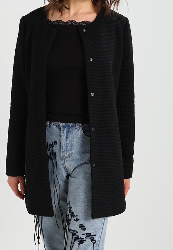 Cardigan largo negro texturizado con escote redondo y botones de presión, combinado con una blusa negra con encaje y vaqueros azul claros con bordado.