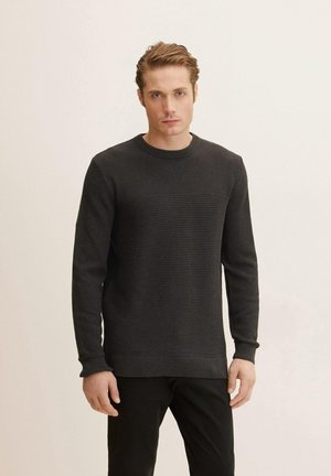 Donker gebreide trui met een gestreepte textuur, ronde halslijn en ribgebreide manchetten. Gecombineerd met een donker broek tegen een neutrale achtergrond.
