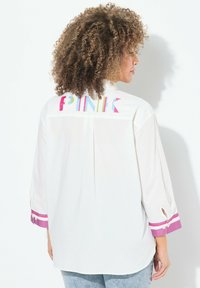 Witte blouse met kleurrijke "PINK" geborduurde tekst op de achterkant, met 3/4 mouwen en roze-witte gestreepte accenten bij de manchetten.