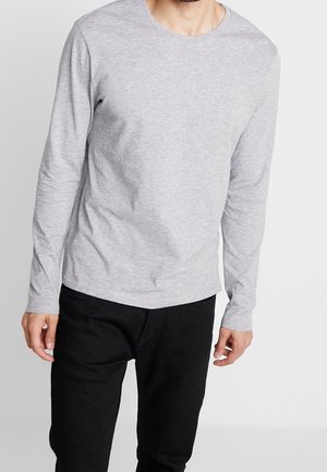 Mann, der ein schlichtes hellgraues Langarmshirt und schwarze Hose trägt, stehend vor einem weißen Hintergrund.
