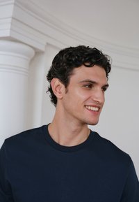 Jeune homme aux cheveux courts, bouclés et foncés, portant une chemise bleu foncé, souriant et regardant vers sa droite sur un fond blanc avec des détails architecturaux.