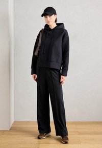 Black hooded sweatshirt with a front pocket, paired with loose black trousers. The model wears a black cap and patterned brown sneakers.