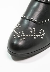Bout de chaussure en cuir noir avec des clous ronds argentés disposés en motifs décoratifs sur fond blanc.
