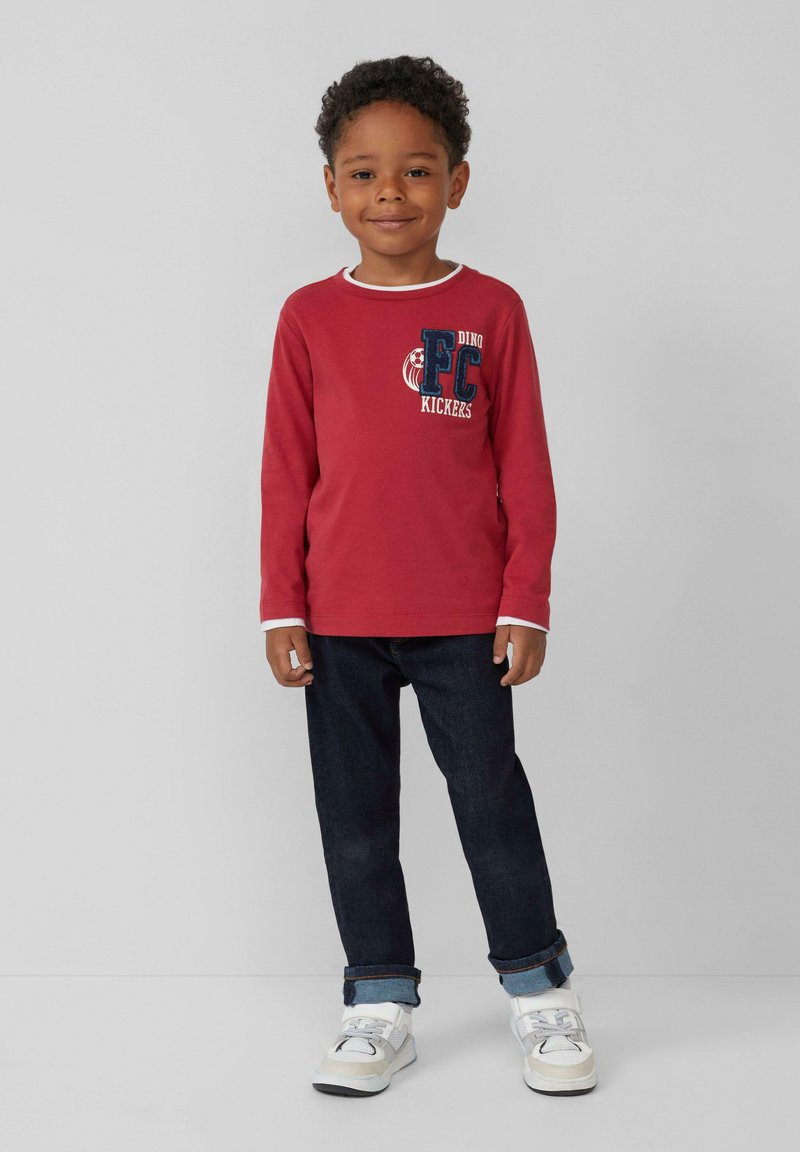 Rotes Langarmhemd mit weißem Saum und blauer gestickter Schrift, kombiniert mit dunklen Jeans-Hosen, die an den Bündchen hochgekrempelt sind, und weißen Sneakern.