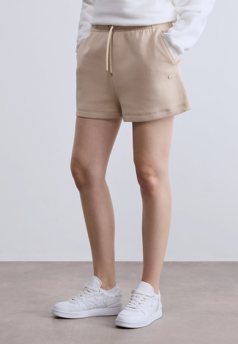 Shorts de survêtement beiges avec une taille élastique et un cordon de serrage, dotés de poches latérales et d'un logo discret sur l'ourlet, fabriqués dans un matériau doux.