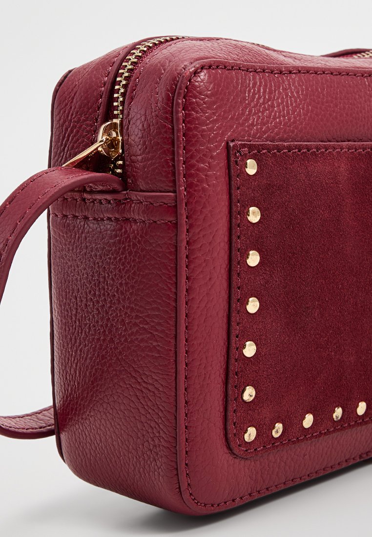 Bordowa skórzana torba o teksturowanym wykończeniu, z zamszową łatką i złotymi ćwiekami. Widok z boku podkreśla detal suwaka i paska.
