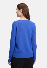 Femme aux longs cheveux bruns portant un pull à manches longues bleu vif et un pantalon plissé noir, de dos sur un fond uni.