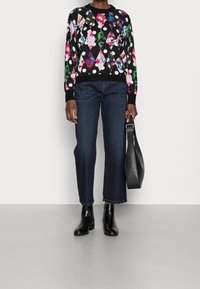 Sort mønstret sweater med blomster- og geometriske design, parret med mørkeblå jeans og sorte ankelstøvler; holder en sort taske.