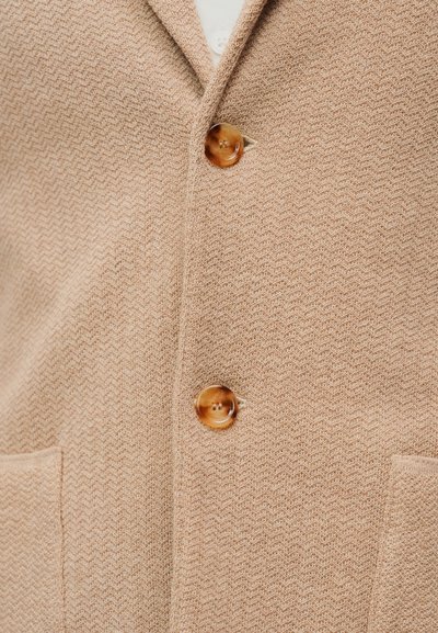 Μπεζ πλεκτή ζακέτα με σχέδιο ψαροκόκαλο, διαθέτει ένα καφέ κουμπί και δύο μπροστινές τσέπες. Ομαλή υφή και εφαρμοστή γραμμή.