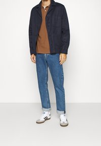 Granatowa kurtka zapinana na guziki over polo w kolorze brązowym, zestawiona z jasnoniebieskimi dżinsami i białymi trampkami w czarne paski. Prosty, casualowy wygląd.