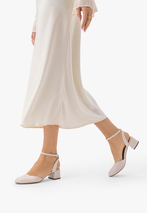 Naine, kes kannab kreemika värvi pahkluu rihmaga plokktallaga kingi ja lendlevat heledat beežikat midi seelikut, seisab valgel taustal.