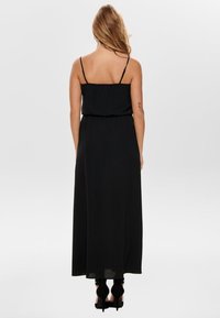 Czarna maxi sukienka z cienkimi regulowanymi ramiączkami, marszczonym wcięciem w talii i gładką teksturą, sięgająca do podłogi. Widać wysokie obcasy.