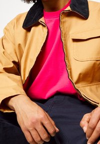 Tan corduroy jacket with black collar, worn over a bright pink t-shirt. Visible pocket detail on the jacket, hands resting on dark blue pants.