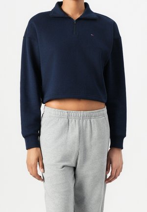 Persona che indossa un maglione cropped a maniche lunghe blu navy con zip corta e pantaloni sportivi elasticizzati in vita di colore grigio chiaro.
