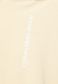 Beżowa tkanina z białym pionowym logo "Calvin Klein Jeans" nadrukowanym na skos wzdłuż powierzchni.