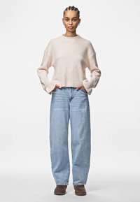 Svijetlo ružičasti skraćeni pulover s rebrastim rubom, uparen s širokim plavim trapericama i smeškim čizmama do gležnjeva, prikazan na jednobojnoj pozadini.