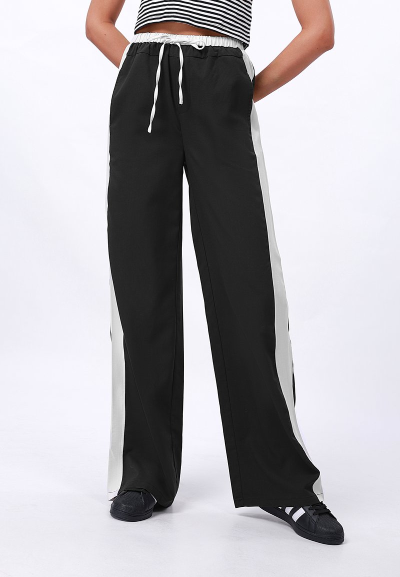 Schwarze weit geschnittene Hose mit elastischem Bund und weißen Seitenstreifen. Hergestellt aus glattem Material, mit einem Kordelzug und einem lässigen Schnitt.
