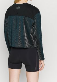 Czarna sportowa kurtka z paskami w kolorze teal, z zamkiem błyskawicznym i logo. W połączeniu z czarnymi szortami kompresyjnymi.