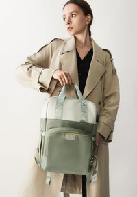 Mochila verde y blanca de forma rectangular, con correas ajustables, un bolsillo frontal y asas con pestañas. Textura suave.