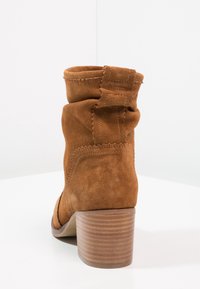 Brune ruskind ankelsko med chunky stakheel og slouch-detail på en hvid overflade med baggrund i hjørnet.