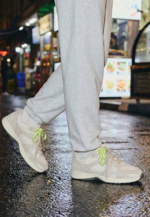 Person wearing light gray sweatpants and beige sneakers with neon yellow laces walking on wet, reflective street pavement at night.