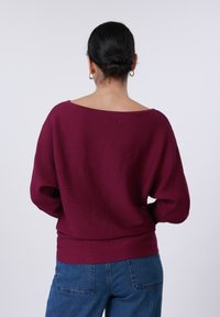 Μπουφάν μπορντό με ριγέ υφή, με φαρδύ λαιμό, μακριά μανίκια και εφαρμοστό τελείωμα. Συνδυασμένο με μπλε τζιν παντελόνια.