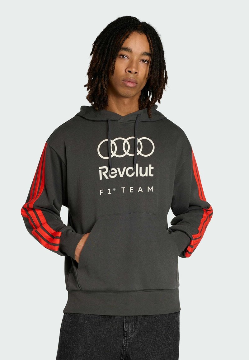 Jeune homme avec des dreadlocks mi-longues portant un sweat à capuche gris foncé avec des rayures rouges sur les manches et le logo "Audi Revolut F1 Team".