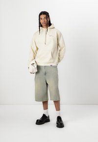 Sweat à capuche beige avec poche frontale et cordon de serrage ; shorts en denim bleu clair avec une apparence décolorée ; chaussures noires avec semelles épaisses.