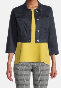 Kvinna klädd i en kort navyjacka med knappar, gul topp och svart-vita rutiga byxor, stående mot en vit bakgrund.