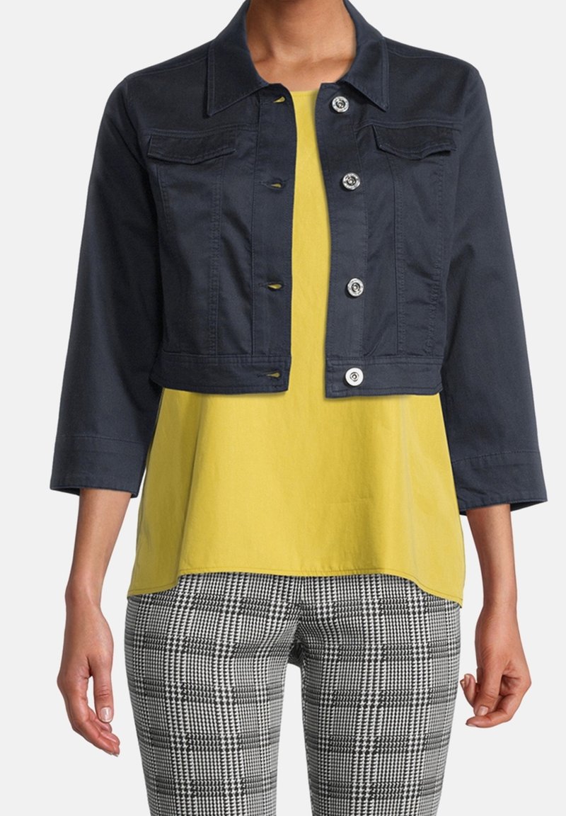 Kvinna klädd i en kort navyjacka med knappar, gul topp och svart-vita rutiga byxor, stående mot en vit bakgrund.