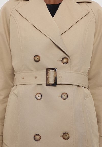 Khaki trench coat with a double-breasted design, large buttons, belted waist, and a notched collar. Smooth fabric with structured pockets.