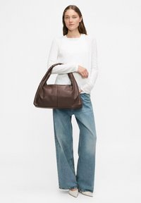 Brązowa skórzana torba na ramię o miękkim, zaokrąglonym kształcie i jednym pasku na ramię. Modelka nosi białą bluzkę i jasnoniebieskie spodnie z szerokimi nogawkami.