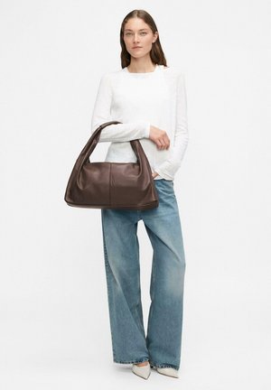 Brązowa skórzana torba na ramię o miękkim, zaokrąglonym kształcie i jednym pasku na ramię. Modelka nosi białą bluzkę i jasnoniebieskie spodnie z szerokimi nogawkami.