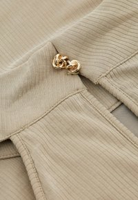 Béžová žebrovaná tkanina s hladkým povrchem, zdobená zlatým kovovým akcentem s propletenými smyčkami. Detailní záběr na švy oděvu.