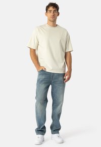 T-shirt a maniche corte color beige chiaro abbinato a jeans blu sbiaditi e sneakers bianche. L'outfit presenta una vestibilità comoda e un design semplice.