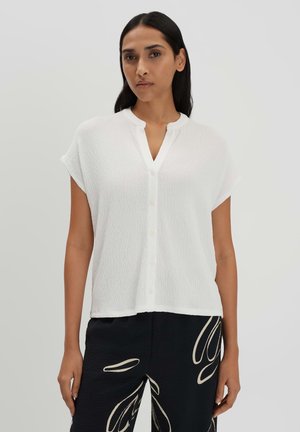 Haut blanc à manches courtes et à boutons, présentant un tissu texturé et un décolleté en V. Associé à un pantalon noir avec un motif clair contrastant.