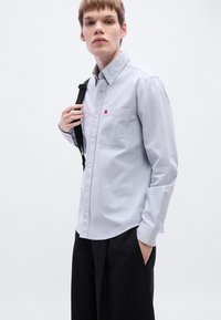 Μακρυμάνικο πουκάμισο ανοιχτού γαλάζιου χρώματος με τσεπάκι στο στήθος και μικρό κόκκινο λογότυπο. Φορεμένο με μαύρο παντελόνι, παρουσιάζοντας έναν μινιμαλιστικό σχεδιασμό.