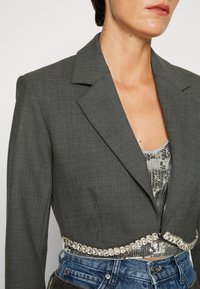 Blazer corto gris con un acabado texturizado y solapas afiladas, que presenta un detalle de cinturón con lentejuelas sobre un top plateado con lentejuelas y pantalones cortos de mezclilla.