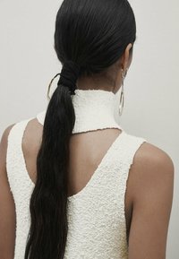 Haut blanc texturé à col montant avec un design ajusté, présentant un dos ouvert. Cheveux longs et noirs attachés avec un chouchou en tissu, et grande boucle d'oreille en argent.