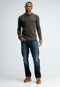 Dunkelgrauer gestrickter Pullover mit Rundhalsausschnitt und kleinem Logo auf der Brust, kombiniert mit hellblauen Denim-Jeans und braunen Wildlederschuhen.