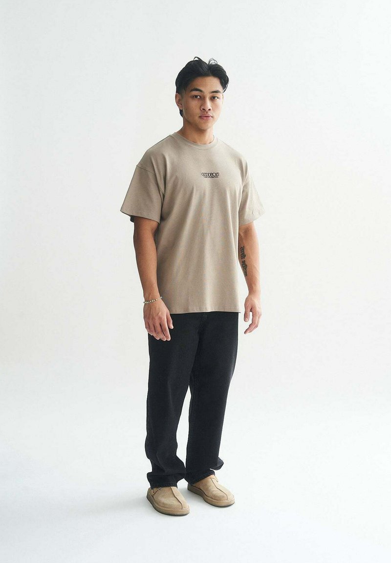 Beige Baumwoll-T-Shirt mit kurzen Ärmeln, schwarze weite Hosen und braune Slipper. Das Modell steht vor einem neutralen Hintergrund mit minimalen Details.
