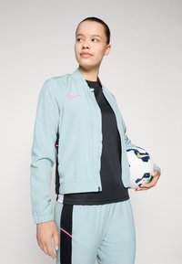 Jachetă Nike de culoare albastru deschis cu un logo roz, top negru și pantaloni asortați, având o textură netedă și manșete elastice. Modelul ține o minge de fotbal.