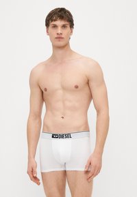 Λευκά ανδρικά εσώρουχα boxer με μαύρη και γκρι ελαστική μέση που φέρει το λογότυπο «DIESEL». Το ύφασμα φαίνεται λείο και αεριζόμενο. Το μοντέλο στέκεται με τα χέρια στο πλευρό.